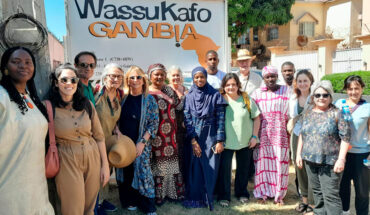 Foto de grup de la delegació que ha viatjat a Gàmbia amb representants de Wassu Gambia Kafo, davant del cartell de l'entitat