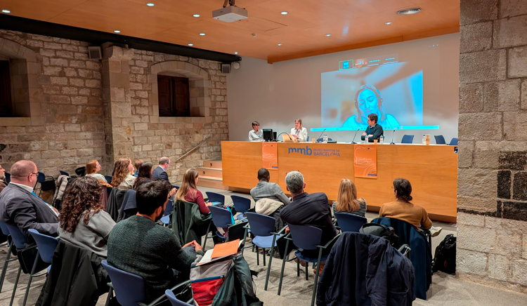 Durant la taula rodona amb dos ponents a dalt la tarima i una pantalla amb una tercera ponent connectada en remot. El públic desquena mirant a l'escenari