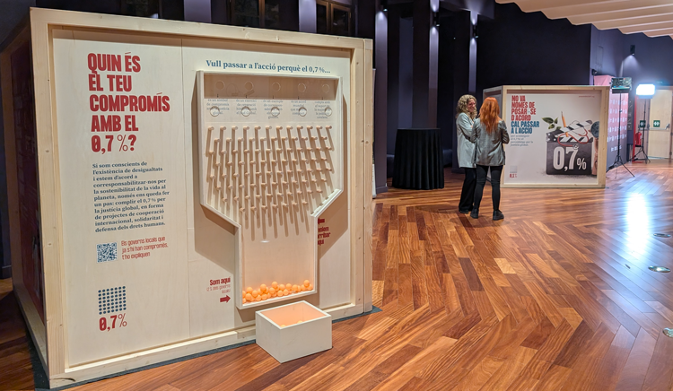 Un cub de fusta sobre l'exposició del 0,7% amb un embut on es poden tirar pilotes de ping-pong, i u segon cub al fons, a una sala de parquet amb dues noies parlant dretes